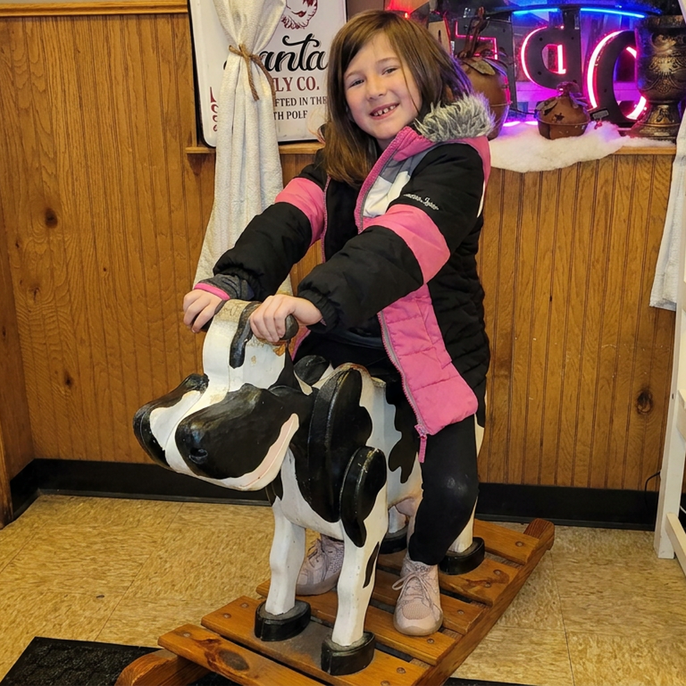 Smiling child in a pink and black winter coat rides a spotted cow sculpture inside a warmly wood-paneled restaurant.