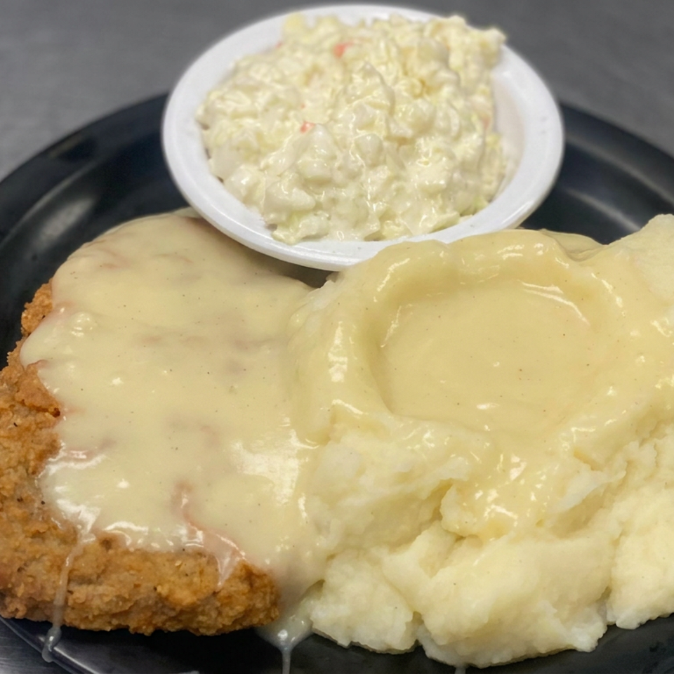 Breaded chicken-fried steak covered in creamy gravy with mashed potatoes and a side of creamy salad on a black plate.