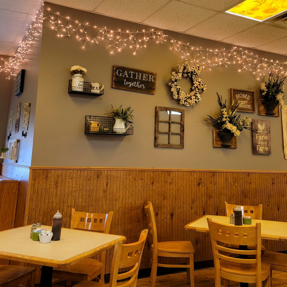 Cozy country-style restaurant dining area with wooden tables, decorative wall signs, floral arrangements, and string lights.