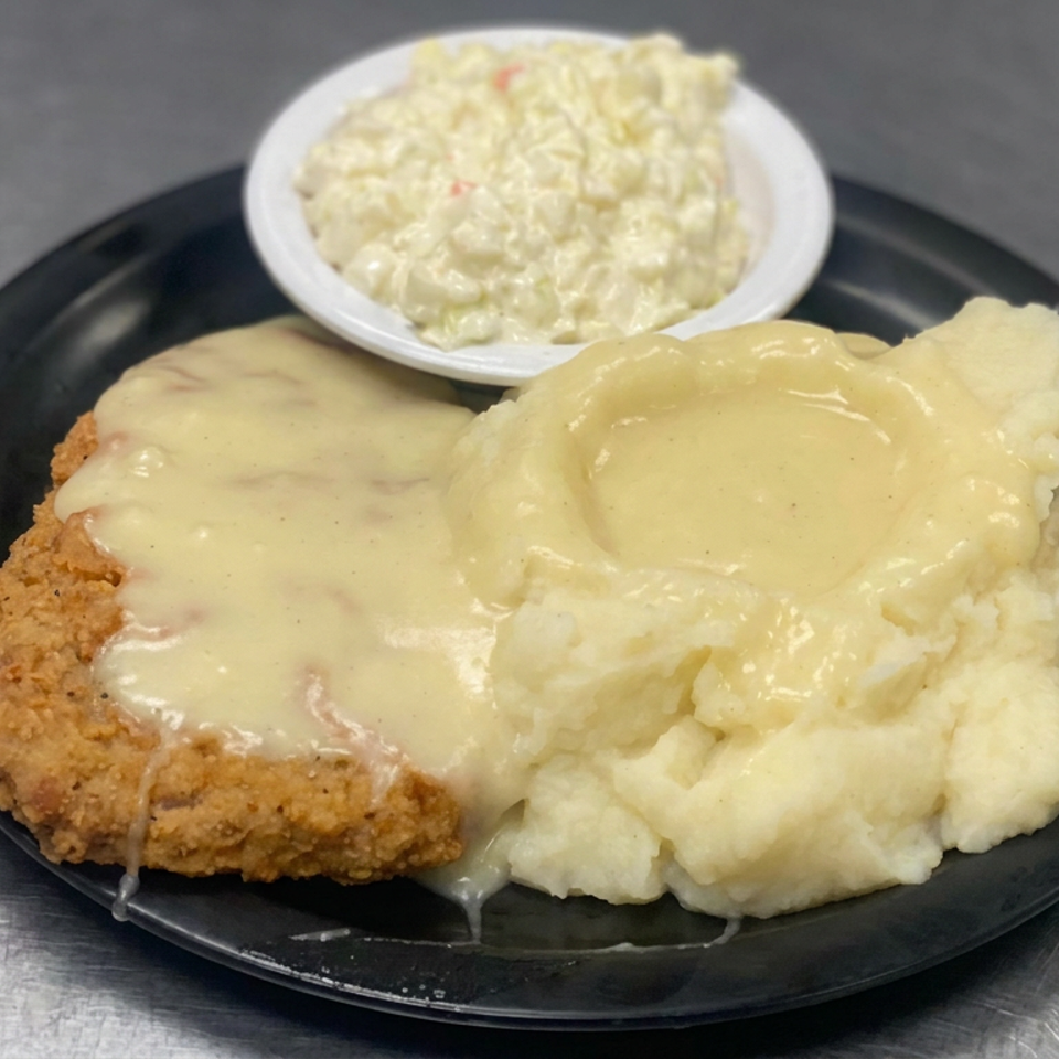 Breaded chicken-fried steak topped with creamy gravy, served with mashed potatoes and a side of creamy salad on a black plate.